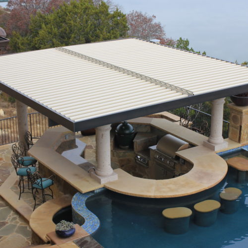 Closed louvers on a motorized pergola provide shade to an outdoor dining area next to the pool in a residential backyard.