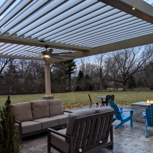 A motorized pergola with a fan attachment covers a backyard seating area in a suburban home. 